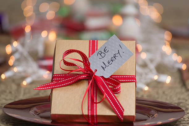 Horizontal close-up photo of gift in gold box with red ribbon at a table set for the holidays. Silver tag with words "BE MERRY". Selective focus with shallow depth of field and lovely background blur.