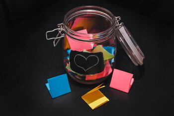 Open Glass Jar with colorful stickers on a black background with white chalk-drawn heart