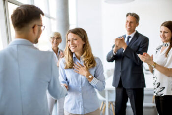 Young business partners making handshake in an office while their team applauding in the background