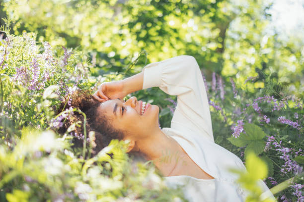 Attractive mixed race girl in white elegant chiffon dress sits among blooming purple flowers. Charming young smiling african woman posing at the camera and enjoying nature. Green trees on background.