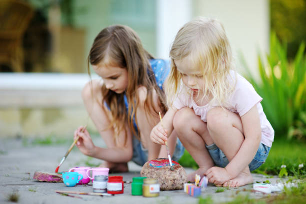 Two cute little sisters painting on stones together on hot summer day